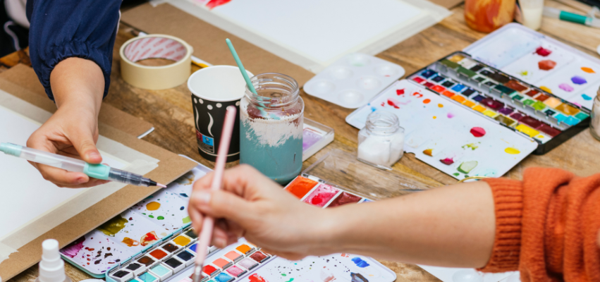 Close-up of hands working with watercolor paints and brushes on a table, surrounded by paint palettes, jars of water, and art supplies, capturing the collaborative atmosphere of a creative workshop.